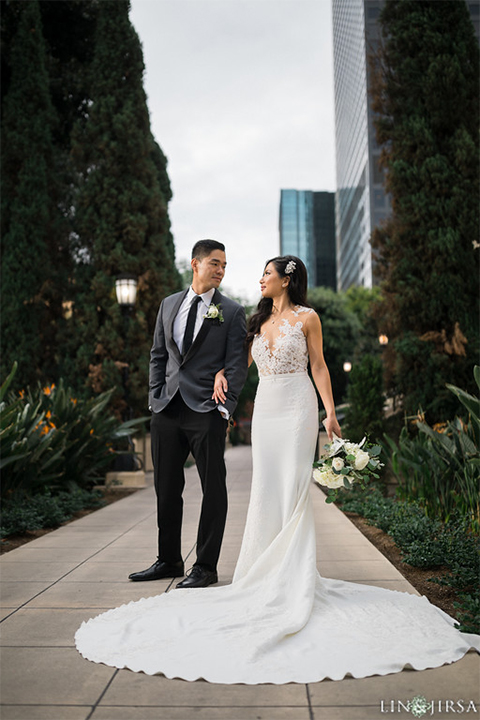 Downtown los angeles wedding bride form fitting lace gown with illusion back and high neckline with crystal hair piece with groom charcoal grey tuxedo with black shawl lapel and white dress shirt with long black skinny tie and white floral boutonniere bride holding white floral bouquet