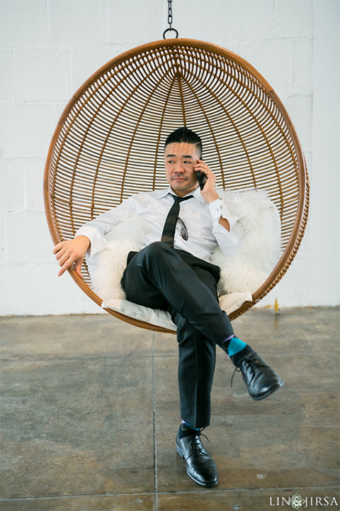 Downtown los angeles wedding groom charcoal grey tuxedo with black shawl lapel and black pants with white dress shirt with long black skinny tie and white floral boutonniere sitting in chair
