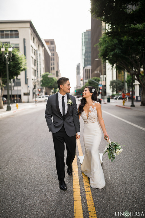 Downtown los angeles wedding bride form fitting lace gown with illusion back and high neckline with crystal hair piece with groom charcoal grey tuxedo with black shawl lapel and white dress shirt with long black skinny tie and white floral boutonniere holding hands and walking in street