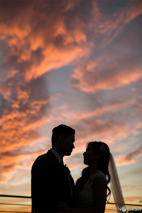 Downtown los angeles wedding bride form fitting lace gown with illusion back and high neckline with crystal hair piece with groom charcoal grey tuxedo with black shawl lapel and white dress shirt with long black skinny tie and white floral boutonniere at sunset