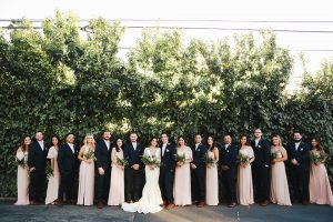Orange county outdoor rustic wedding bride form fitting lace gown with sleeves and plunging neckline with groom navy blue notch lapel suit with white dress shirt and blush pink bow tie with matching pocket square and whit and green floral boutonniere with bridesmaids long blush pink dresses and groomsmen navy blue suits with blush pink bow ties