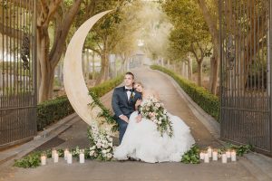 California wedding shoot at enchanted forest bride ball gown with high illusion neckline and lace sleeves with ruffled skirt and open back design with groom navy blue shawl lapel tuxedo with matching vest and white dress shirt with black bow tie and white floral boutonniere sitting on crescent moon decor with flowers and candles on ground bride holding white and green floral bridal bouquet