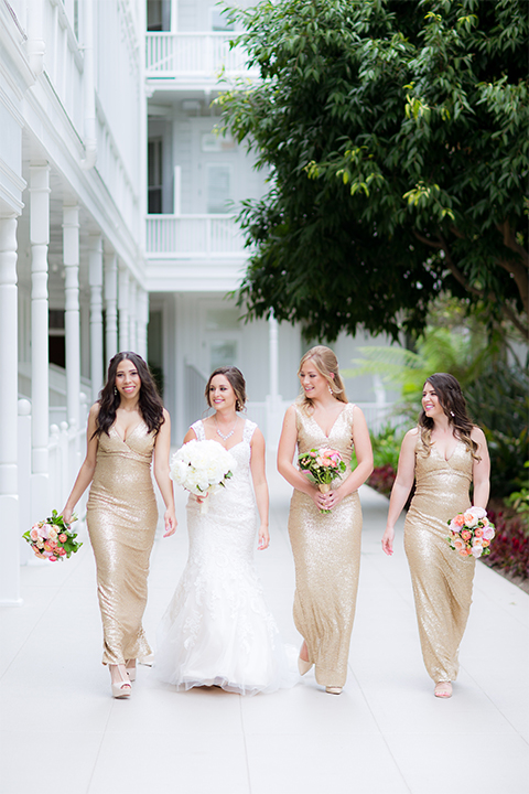 San diego wedding at the crossings carlsbad bride form fitting lace gown with thin straps and ruffled skirt with sweetheart neckline holding white floral bridal bouquet with bridesmaids long gold sequined dresses with high necklines and white and pink floral bridal bouquets walking