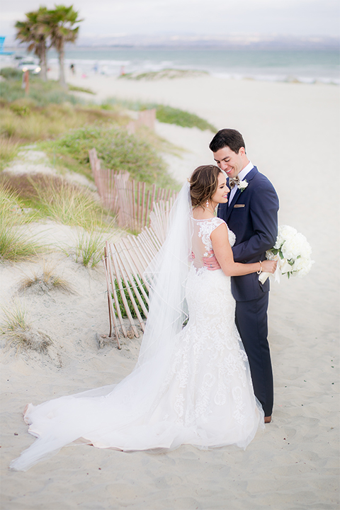 San diego wedding at the crossings carlsbad bride form fitting lace gown with thin straps and ruffled skirt with sweetheart neckline and long veil with groom cobalt blue suit with white dress shirt and gold bow tie with matching pocket square and white floral boutonniere hugging on the beach bride holding white floral bridal bouquet