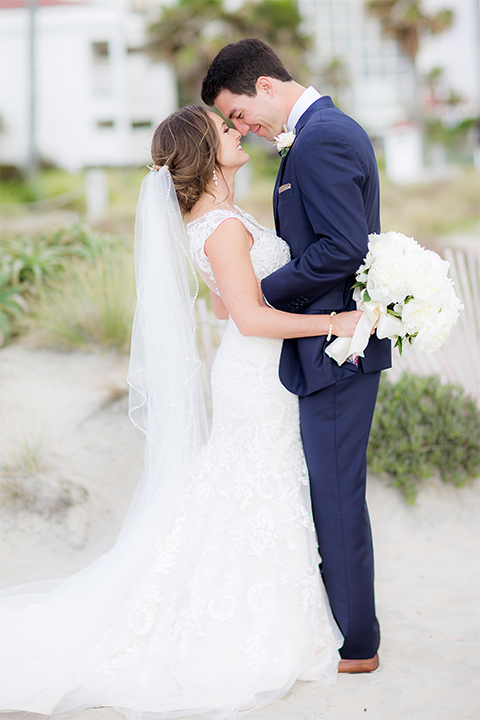 San diego wedding at the crossings carlsbad bride form fitting lace gown with thin straps and ruffled skirt with sweetheart neckline and long veil with groom cobalt blue suit with white dress shirt and gold bow tie with matching pocket square and white floral boutonniere hugging on the beach bride holding white floral bridal bouquet