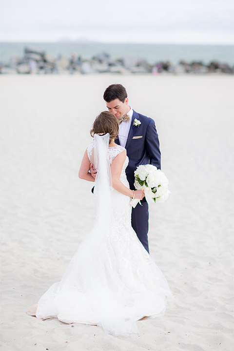 San diego wedding at the crossings carlsbad bride form fitting lace gown with thin straps and ruffled skirt with sweetheart neckline and long veil with groom cobalt blue suit with white dress shirt and gold bow tie with matching pocket square and white floral boutonniere hugging on the beach bride holding white floral bridal bouquet
