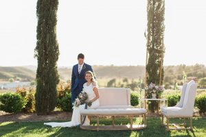 Temecula outdoor wedding shoot at avensole winery bride form fitting gown with lace detail on short sleeves and high neckline with hair in side braid with groom navy blue suit with matching vest and white dress shirt with long floral tie and green and blue floral boutonniere standing next to bride sitting on white couch with purple and green floral bridal bouquet