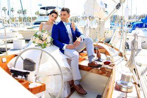 Los angeles wedding shoot at LA yacht club bride ball gown with beaded bodice and thin straps with open back design and high neckline with groom cobalt blue notch lapel suit with light grey pants and matching vest with white dress shirt and light blue striped bow tie sitting on boat bride holding white and green floral bridal bouquet