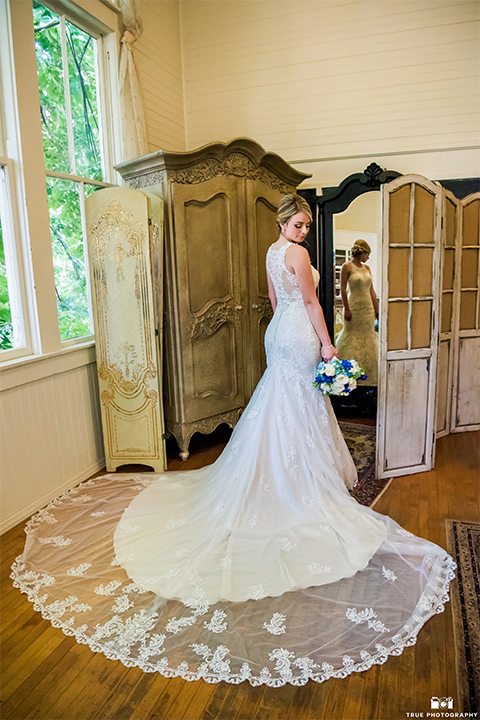 San diego wedding at green gables estate bride form fitting lace gown with a long train and high illusion neckline with long veil holding white and blue floral bridal bouquet