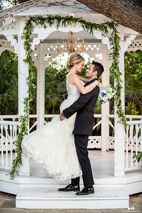 San diego wedding at green gables estate bride form fitting lace gown with a long train and high illusion neckline with long veil and groom black shawl lapel tuxedo with matching vest and white dress shirt with long black skinny tie and white floral boutonniere hugging and smiling