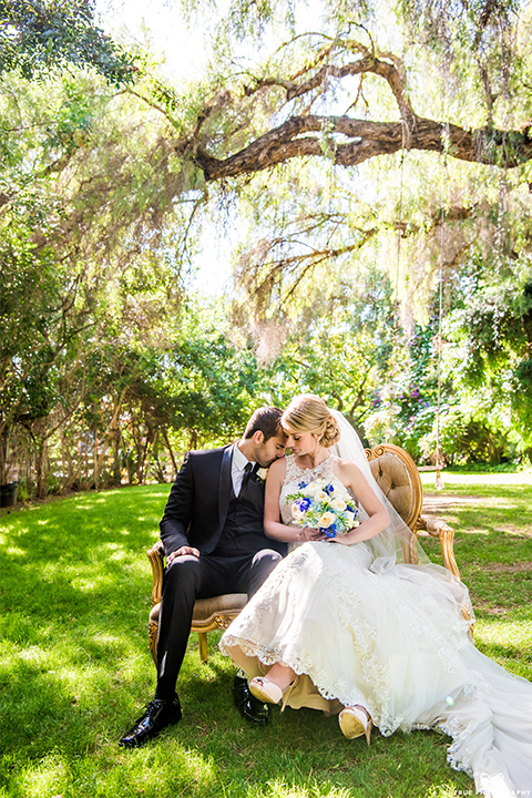 San diego wedding at green gables estate bride form fitting lace gown with a long train and high illusion neckline with long veil and groom black shawl lapel tuxedo with matching vest and white dress shirt with long black skinny tie and white floral boutonniere hugging and bride holding white and blue floral bridal bouquet