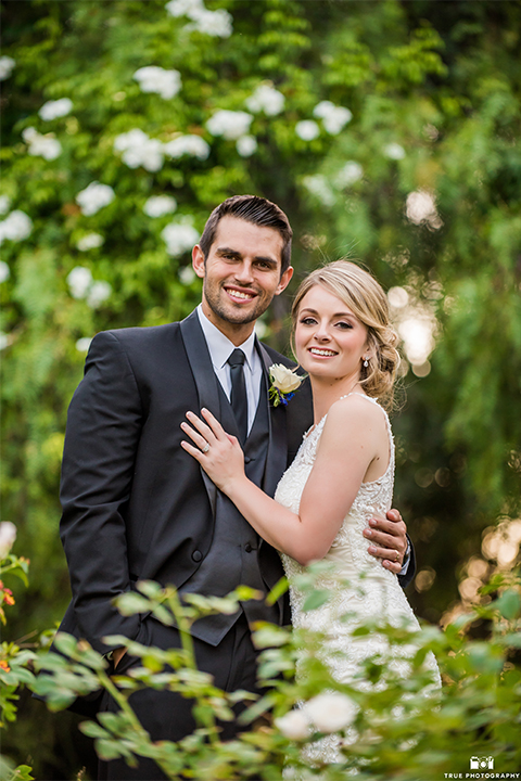 San diego wedding at green gables estate bride form fitting lace gown with a long train and high illusion neckline with long veil and groom black shawl lapel tuxedo with matching vest and white dress shirt with long black skinny tie and white floral boutonniere hugging