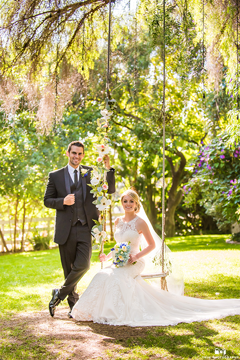 San diego wedding at green gables estate bride form fitting lace gown with a long train and high illusion neckline with long veil and groom black shawl lapel tuxedo with matching vest and white dress shirt with long black skinny tie and white floral boutonniere bride sitting on swing and groom standing