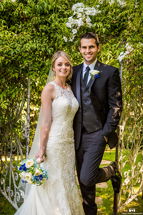 San diego wedding at green gables estate bride form fitting lace gown with a long train and high illusion neckline with long veil and groom black shawl lapel tuxedo with matching vest and white dress shirt with long black skinny tie and white floral boutonniere hugging and bride holding white and blue floral bridal bouquet