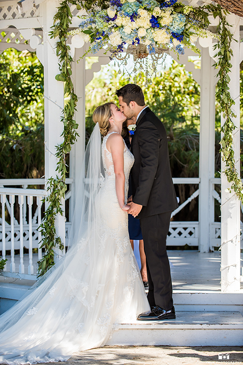 San diego wedding at green gables estate bride form fitting lace gown with a long train and high illusion neckline with long veil and groom black shawl lapel tuxedo with matching vest and white dress shirt with long black skinny tie and white floral boutonniere holding hands and kissing during ceremony
