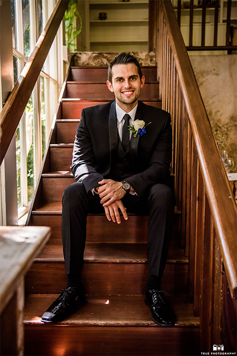 San diego wedding at green gables estate groom black shawl lapel tuxedo with matching vest and white dress shirt with long black skinny tie and white floral boutonniere sitting on stair case