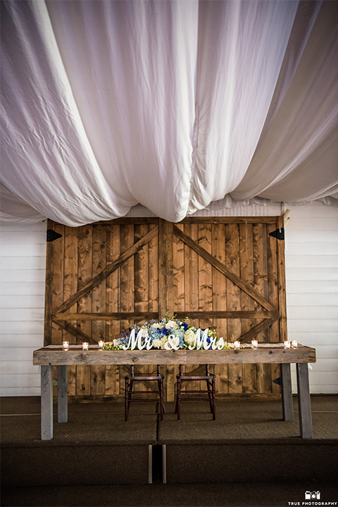 San diego wedding at green gables estate table set up light brown wood table with white and blue flower decor and white place settings with blue napkins and white candles with white draping on ceiling and hanging chandeliers