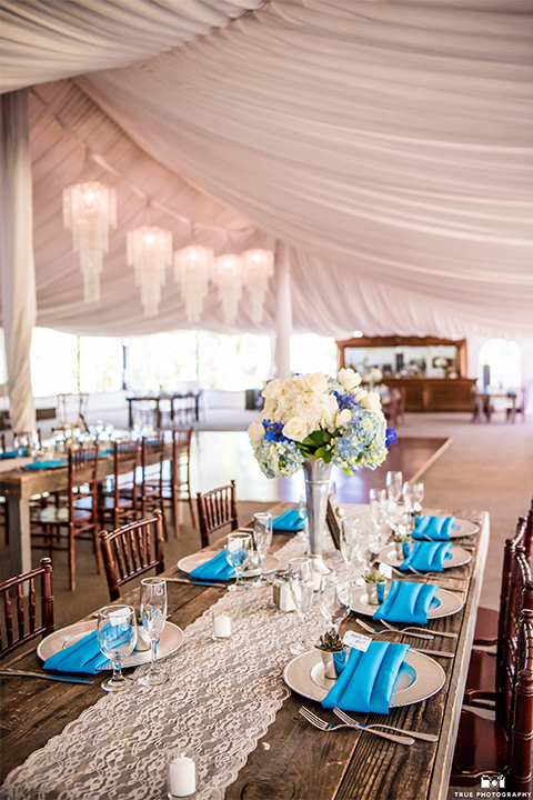 San diego wedding at green gables estate table set up light brown wood table with white and blue flower decor and white place settings with blue napkins and white candles with white draping on ceiling and hanging chandeliers