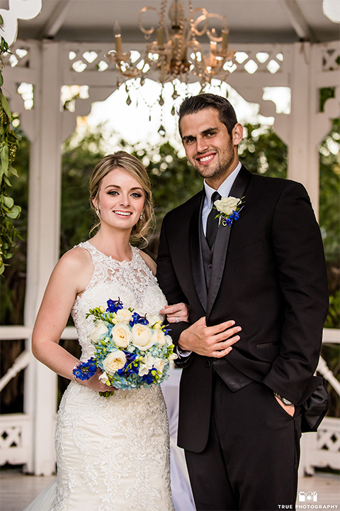 San diego wedding at green gables estate bride form fitting lace gown with a long train and high illusion neckline with long veil and groom black shawl lapel tuxedo with matching vest and white dress shirt with long black skinny tie and white floral boutonniere hugging and bride holding white and blue floral bridal bouquet