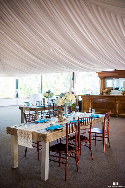 San diego wedding at green gables estate table set up light brown wood table with white and blue flower decor and white place settings with blue napkins and white candles with white draping on ceiling and hanging chandeliers
