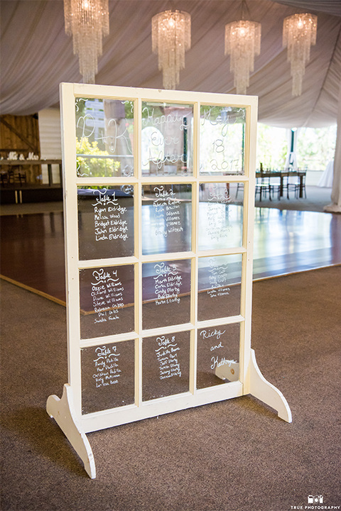 San diego wedding at green gables estate table set up light brown wood table with white and blue flower decor and white place settings with blue napkins and white candles with white draping on ceiling and hanging chandeliers with window pane seating guest list decor