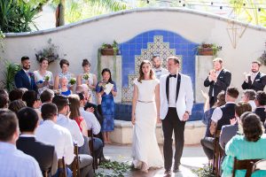 Orange county outdoor wedding at the green parrot villa bride form fitting white gown with a high neckline and crystal belt with short veil and groom white tuxedo jacket with black shawl lapel and black pants with white dress shirt and black bow tie holding hands and walking back down the aisle after ceremony