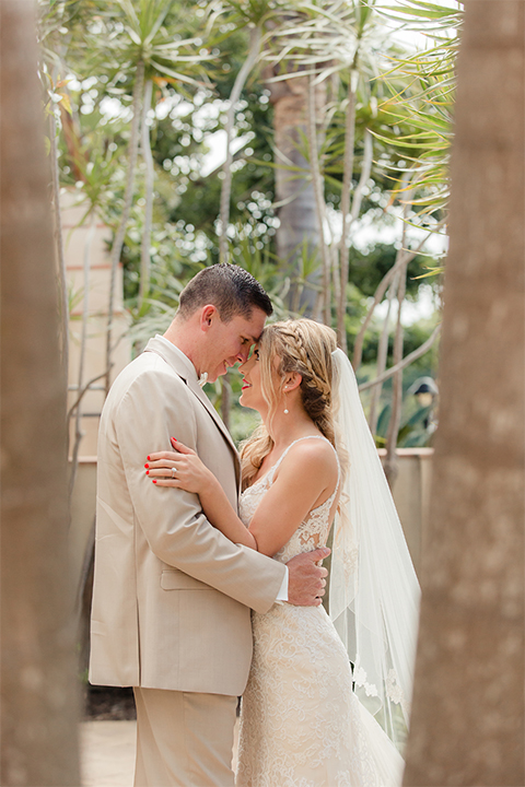 Los angeles wedding at los verdes golf club bride form fitting gown with lace detail and low back design with thin straps and long veil with groom tan notch lapel suit with matching vest and white dress shirt with matching tan bow tie and white pocket square with white and red floral boutonniere hugging