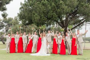 Los angeles wedding at los verdes golf club bride form fitting gown with lace detail and low back design with thin straps and long veil with groom tan notch lapel suit with matching vest and white dress shirt with matching tan bow tie and white pocket square with white and red floral boutonniere with wedding party bridesmaids long red dresses and groomsmen tan suits with red bow ties cheering