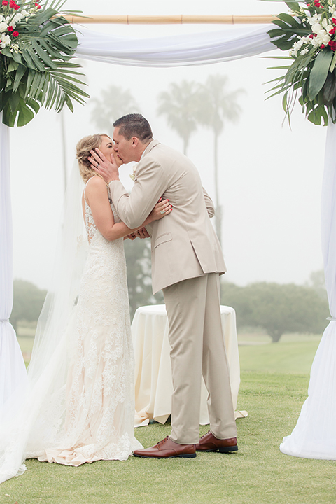 Los angeles wedding at los verdes golf club bride form fitting gown with lace detail and low back design with thin straps and long veil with groom tan notch lapel suit with matching vest and white dress shirt with matching tan bow tie and white pocket square with white and red floral boutonniere kissing during ceremony