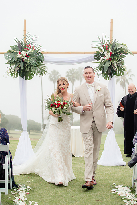 Los angeles wedding at los verdes golf club bride form fitting gown with lace detail and low back design with thin straps and long veil with groom tan notch lapel suit with matching vest and white dress shirt with matching tan bow tie and white pocket square with white and red floral boutonniere walking back down the aisle after ceremony