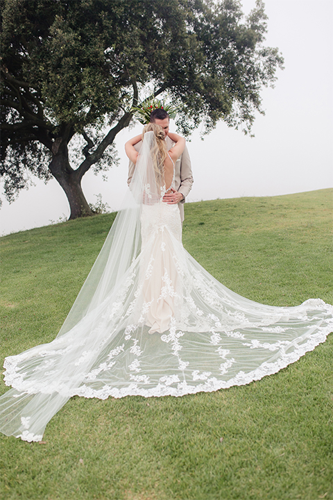 Los angeles wedding at los verdes golf club bride form fitting gown with lace detail and low back design with thin straps and long veil with groom tan notch lapel suit with matching vest and white dress shirt with matching tan bow tie and white pocket square with white and red floral boutonniere hugging and kissing