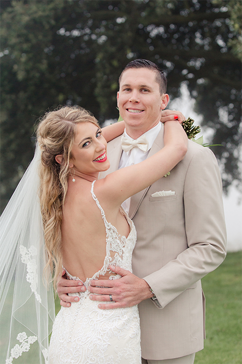Los angeles wedding at los verdes golf club bride form fitting gown with lace detail and low back design with thin straps and long veil with groom tan notch lapel suit with matching vest and white dress shirt with matching tan bow tie and white pocket square with white and red floral boutonniere hugging