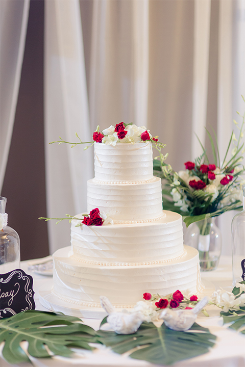 Los verdes golf club wedding table set up white table linens with dark chairs and white and green and red flower centerpiece decor with wine glasses and table numbers with wedding cake four tier white wedding cake with red and white flowers on white table linen