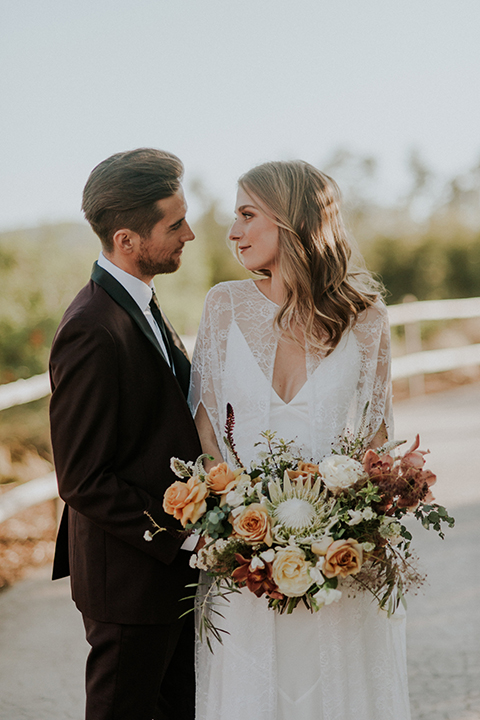 San diego outdoor wedding shoot at the san diego zoo and safari park bride white chiffon gown with lace sleeves and thin straps with plunging neckline and groom burgundy shawl lapel tuxedo with white dress shirt and long black skinny tie and black shoes hugging and bride holding white and orange floral bridal bouquet