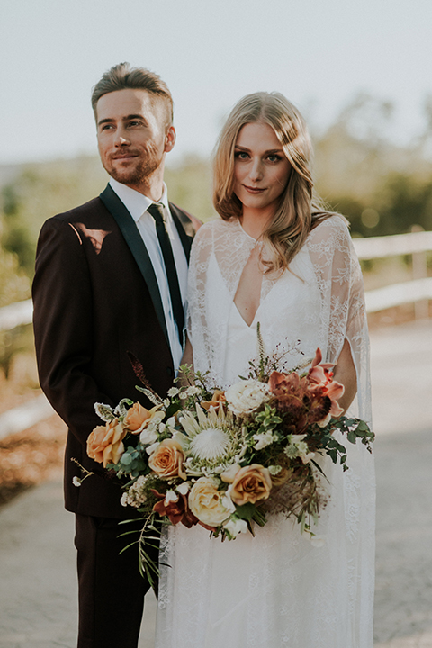 San diego outdoor wedding shoot at the san diego zoo and safari park bride white chiffon gown with lace sleeves and thin straps with plunging neckline and groom burgundy shawl lapel tuxedo with white dress shirt and long black skinny tie and black shoes hugging and bride holding white and orange floral bridal bouquet