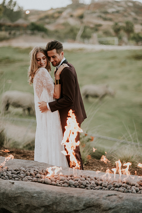 San diego outdoor wedding shoot at the san diego zoo and safari park bride white chiffon gown with lace sleeves and thin straps with plunging neckline and groom burgundy shawl lapel tuxedo with white dress shirt and long black skinny tie and black shoes standing and hugging by fire