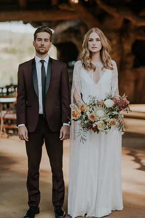 San diego outdoor wedding shoot at the san diego zoo and safari park bride white chiffon gown with lace sleeves and thin straps with plunging neckline and groom burgundy shawl lapel tuxedo with white dress shirt and long black skinny tie and black shoes standing holding white and orange floral bridal bouquet