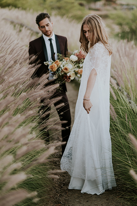 San diego outdoor wedding shoot at the san diego zoo and safari park bride white chiffon gown with lace sleeves and thin straps with plunging neckline and groom burgundy shawl lapel tuxedo with white dress shirt and long black skinny tie and black shoes standing and bride holding white and orange floral bridal bouquet