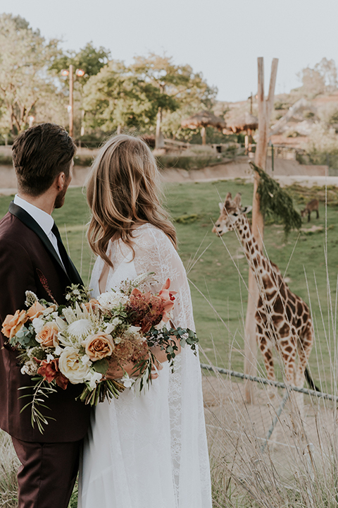 San diego outdoor wedding shoot at the san diego zoo and safari park bride white chiffon gown with lace sleeves and thin straps with plunging neckline and groom burgundy shawl lapel tuxedo with white dress shirt and long black skinny tie and black shoes hugging and bride holding white and orange floral bridal bouquet