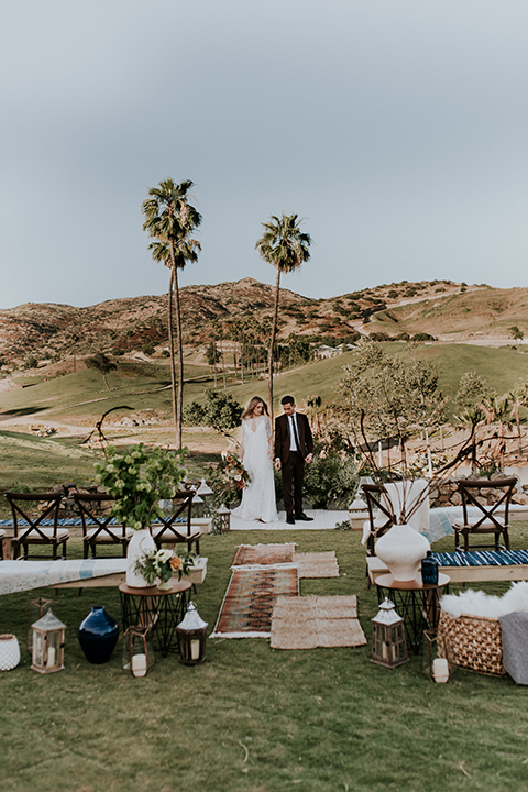 San diego outdoor wedding shoot at the san diego zoo and safari park bride white chiffon gown with lace sleeves and thin straps with plunging neckline and groom burgundy shawl lapel tuxedo with white dress shirt and long black skinny tie and black shoes standing and holding hands during ceremony