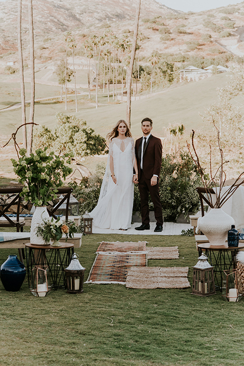 San diego outdoor wedding shoot at the san diego zoo and safari park bride white chiffon gown with lace sleeves and thin straps with plunging neckline and groom burgundy shawl lapel tuxedo with white dress shirt and long black skinny tie and black shoes standing and holding hands during ceremony