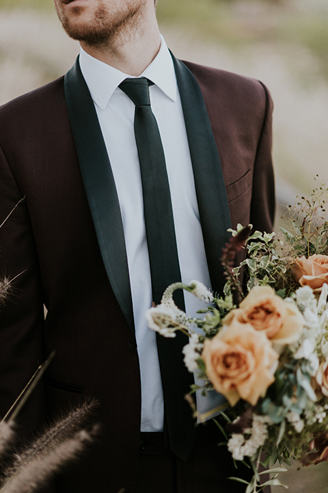 San diego outdoor wedding shoot at the san diego zoo and safari park groom burgundy tuxedo with black shawl lapel and white dress shirt with long black skinny tie and black shoes holding white and orange floral bridal bouquet close up