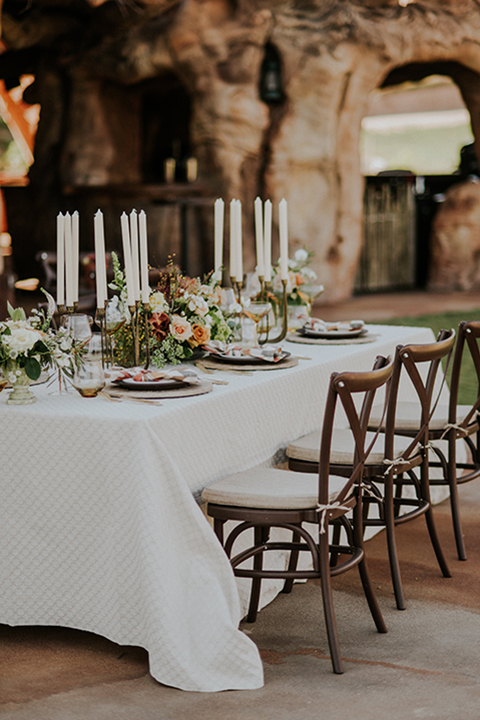 San diego outdoor wedding shoot at the san diego zoo and safari park table set up with white table linens and brown wood chairs with brown and white place settings and multi colored napkins with red place card and white and orange flower centerpiece decor with gold silverware and tall white candles