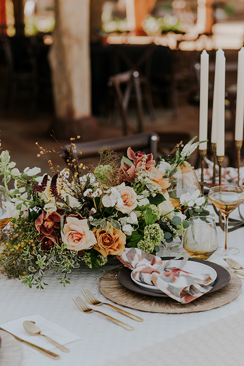 San diego outdoor wedding shoot at the san diego zoo and safari park table set up with white table linens and brown wood chairs with brown and white place settings and multi colored napkins with red place card and white and orange flower centerpiece decor with gold silverware and tall white candles