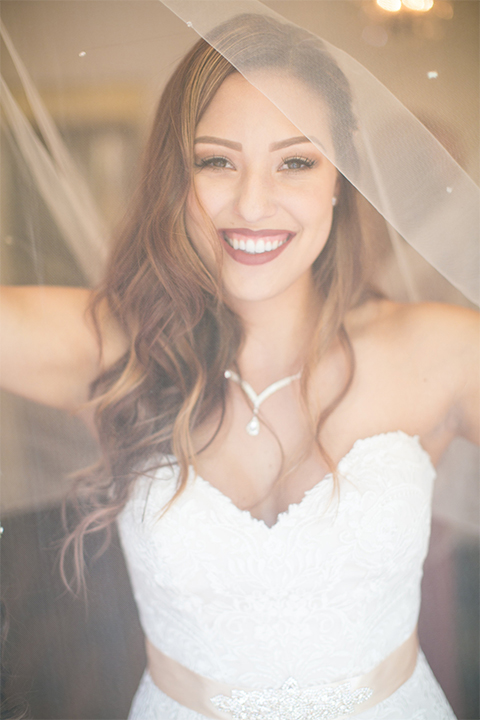 Temecula outdoor rustic wedding at lake oak meadows bride form fitting strapless gown with sweetheart neckline and lace detail with long veil with necklace holding white and red floral bridal bouquet with red ribbon decor