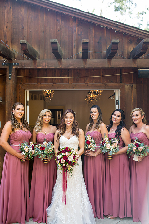 Temecula outdoor rustic wedding at lake oak meadows bride form fitting strapless gown with sweetheart neckline and lace detail with long veil with necklace holding white and red floral bridal bouquet with red ribbon decor with bridesmaids long dusty pink dresses with red and white floral bouquets