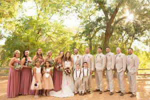Temecula outdoor rustic wedding at lake oak meadows bride form fitting strapless gown with sweetheart neckline and lace detail with long veil and groom tan notch lapel suit with matching vest and white dress shirt and long white tie with red floral boutonniere with wedding party bridesmaids long dusty pink dresses with white and red floral bouquets and groomsmen tan suits with long ties