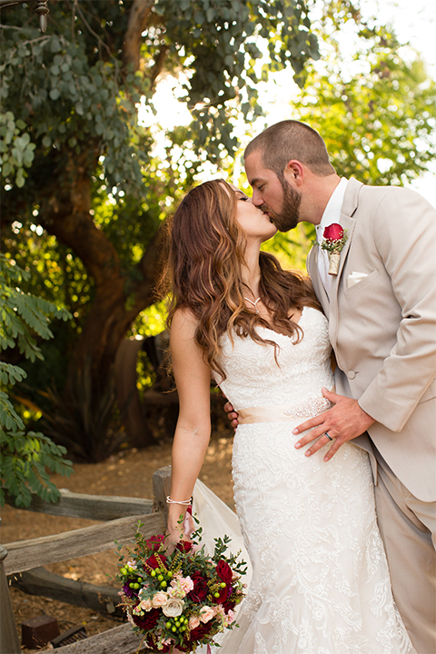 Temecula outdoor rustic wedding at lake oak meadows bride form fitting strapless gown with sweetheart neckline and lace detail with long veil and groom tan notch lapel suit with matching vest and white dress shirt and long white tie with red floral boutonniere kissing and bride holding white and red floral bridal bouquet with red ribbon decor