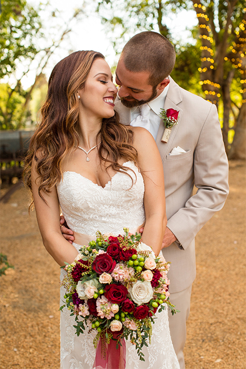 Temecula outdoor rustic wedding at lake oak meadows bride form fitting strapless gown with sweetheart neckline and lace detail with long veil and groom tan notch lapel suit with matching vest and white dress shirt and long white tie with red floral boutonniere hugging and bride holding white and red floral bridal bouquet with red ribbon decor