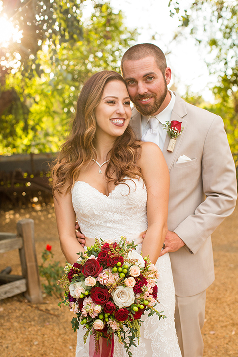 Temecula outdoor rustic wedding at lake oak meadows bride form fitting strapless gown with sweetheart neckline and lace detail with long veil and groom tan notch lapel suit with matching vest and white dress shirt and long white tie with red floral boutonniere hugging and holding white and red floral bridal bouquet with red ribbon decor